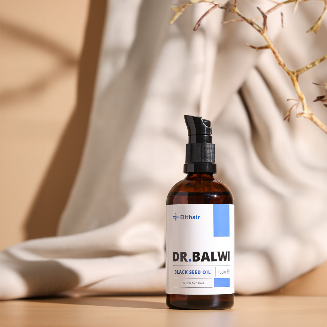 Auf einem beigen Untergrund steht eine braune Pumpflasche aus Glas mit der Aufschrift „Premium Schwarzkümmelöl“ von shop.elithairtransplant, ideal für die Haarpflege, umgeben von weichem Stoff und getrockneten Pflanzenzweigen in sanfter, natürlicher Umgebung.