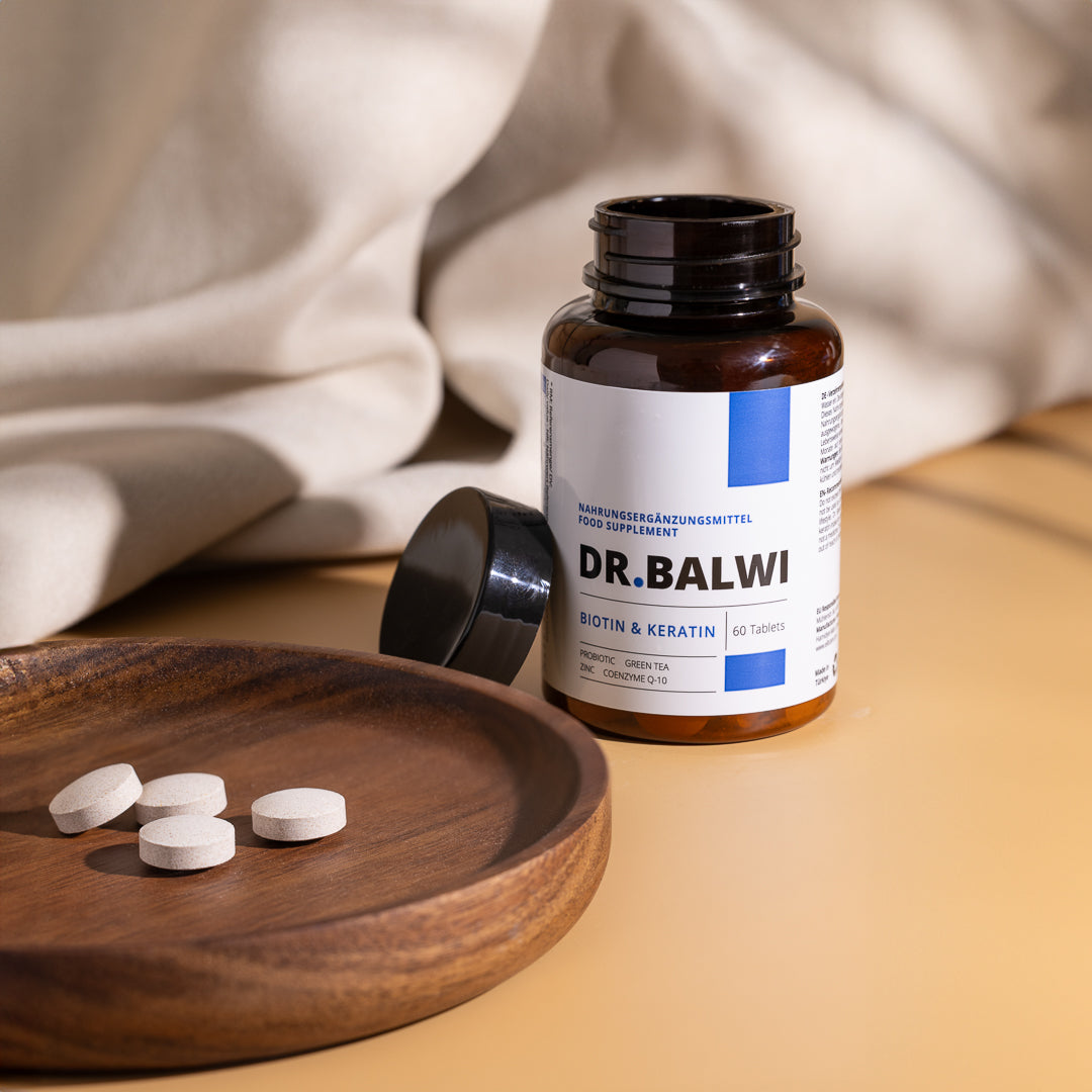 Eine offene braune Flasche aus dem Dr. Balwi Hair Boost Set von shop.elithairtransplant steht auf einer beigen Oberfläche, mit vier runden Tabletten auf einer Holzschale und weichem, hellem Stoff im Hintergrund.