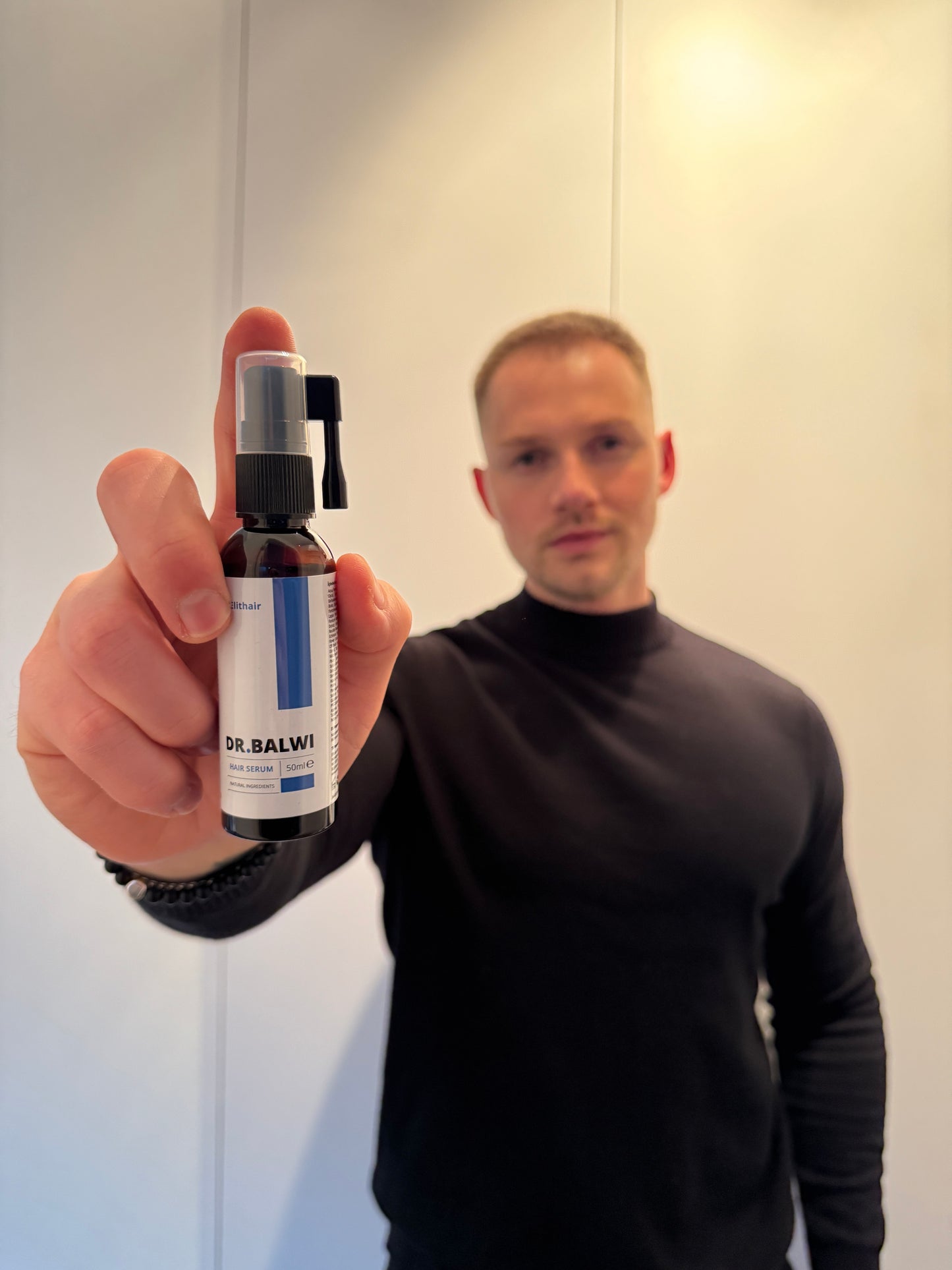 Ein Mann im schwarzen Hemd hält eine Flasche Dr. Balwi Hair Serum von shop.elithairtransplant nah an die Kamera und hebt das hochkonzentrierte Serum für Haarwachstum hervor, während sein Gesicht im Hintergrund leicht unscharf erscheint.