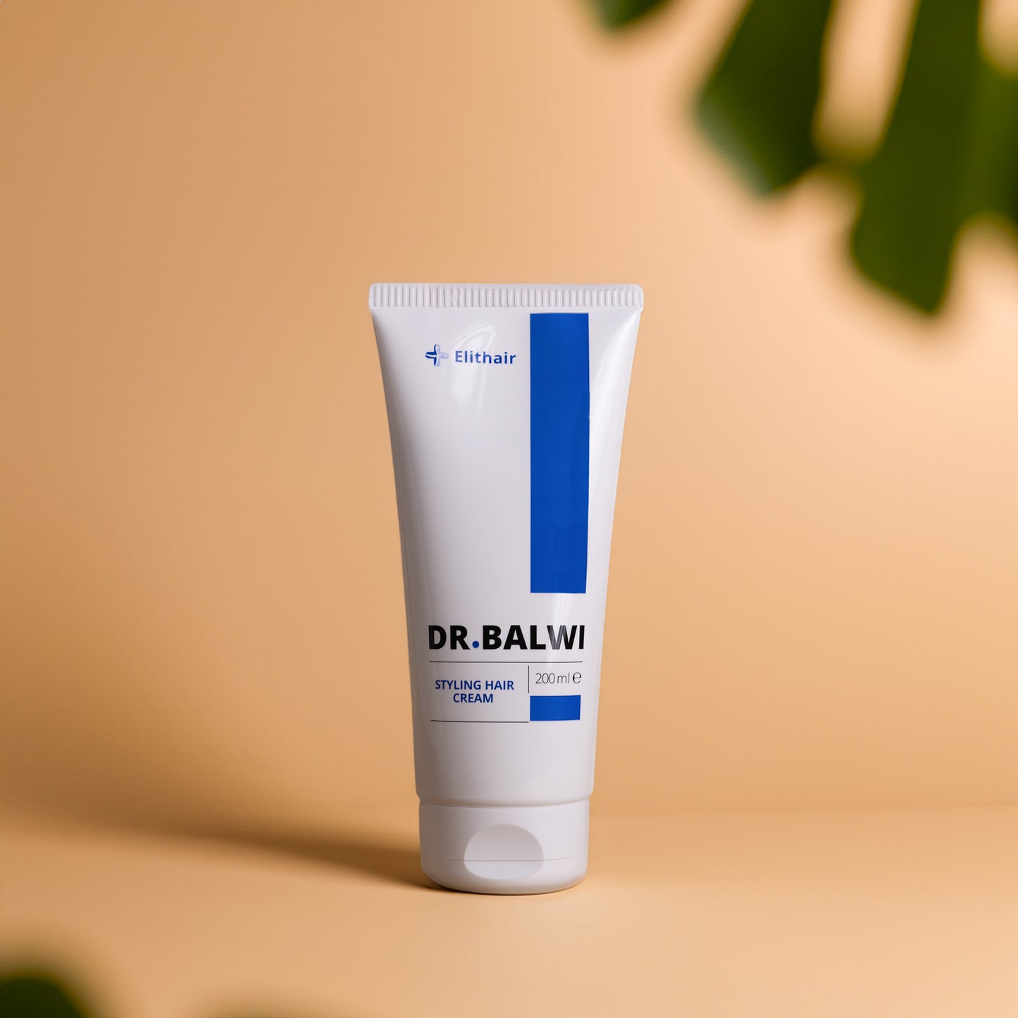 Eine weiße Tube Dr. Balwi Styling & Repair Creme von shop.elithairtransplant mit blauen Akzenten steht aufrecht auf einem beigen Untergrund und ist mit Avocadoöl und Arganöl versehen. Das 200-ml-Produkt ist mit verschwommenen grünen Blättern im Vordergrund abgebildet.