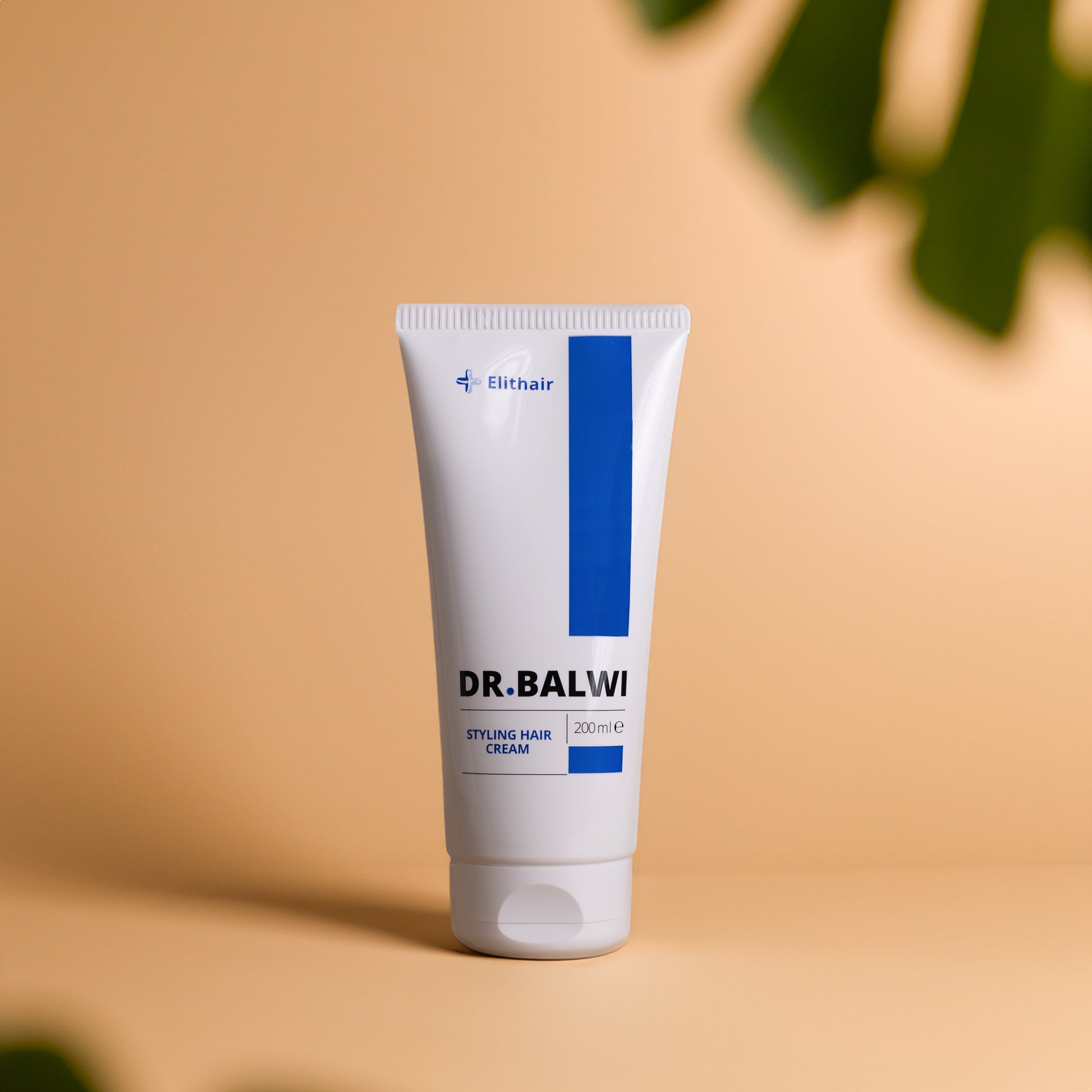 Eine weiße Tube Dr. Balwi Styling & Repair Creme von shop.elithairtransplant mit blauen Akzenten steht aufrecht auf einem beigen Untergrund und ist mit Avocadoöl und Arganöl versehen. Das 200-ml-Produkt ist mit verschwommenen grünen Blättern im Vordergrund abgebildet.