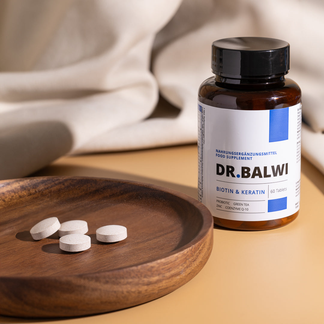 Eine braune Flasche mit der Aufschrift „Biotin & Keratin Tabletten“ von shop.elithairtransplant steht auf einem beigen Untergrund neben einem Holzteller mit vier runden Tabletten. Der Hintergrund besteht aus weichem, hellem Stoff.