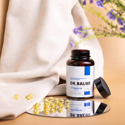 Eine Flasche Vitamin D 1000 IE Softgels von shop.elithairtransplant steht auf einer Spiegelfläche, daneben gelbe Kapseln. Ein beiges Tuch und violette Blumen bilden einen weichen, natürlichen Hintergrund – ideal zur Unterstützung des Vitamin-D- und Immunsystembedarfs.