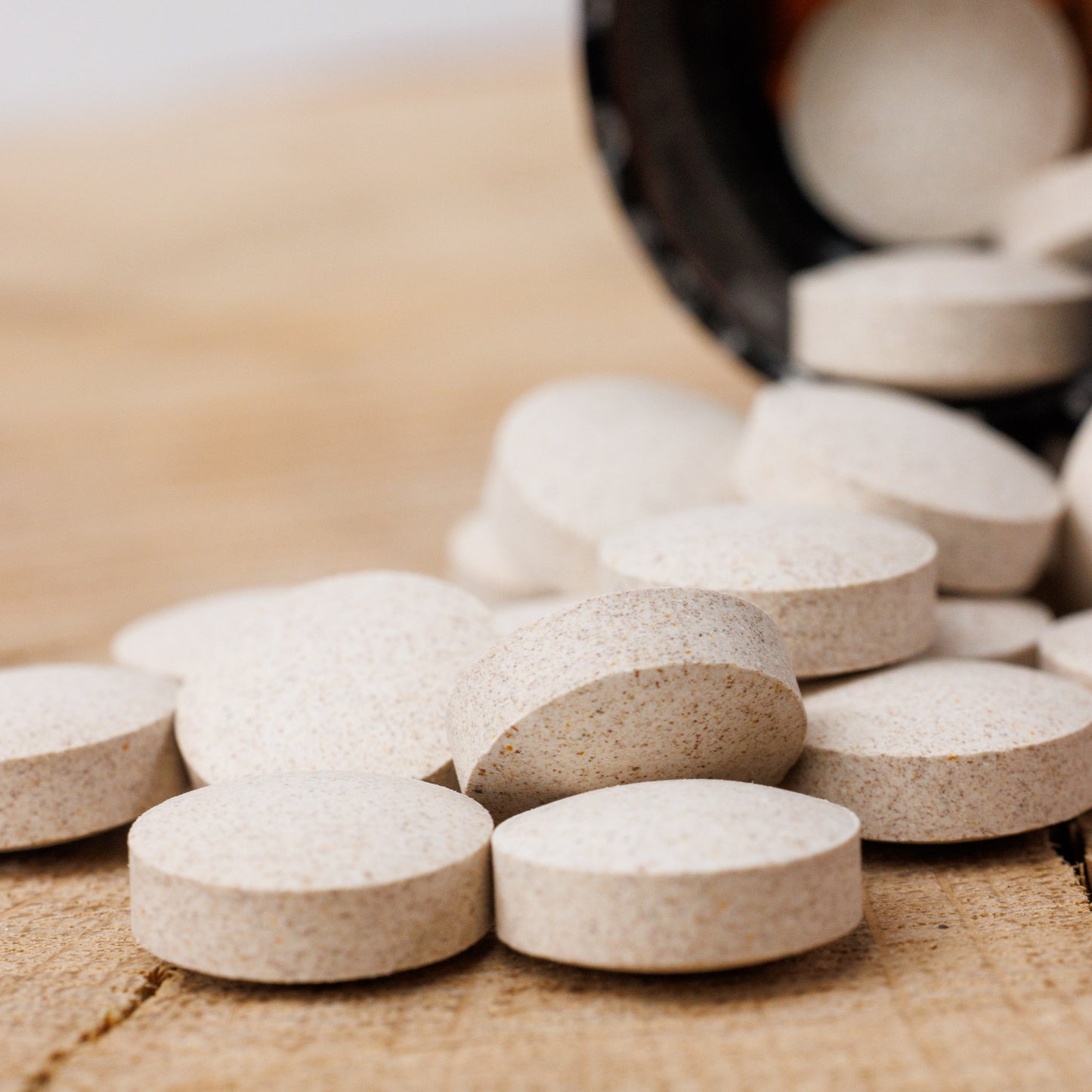 Eine Nahaufnahme runder, beiger Biotin- und Keratintabletten von shop.elithairtransplant, die auf Holz verstreut sind, während im Hintergrund weitere Tabletten aus einer offenen dunklen Flasche quellen.