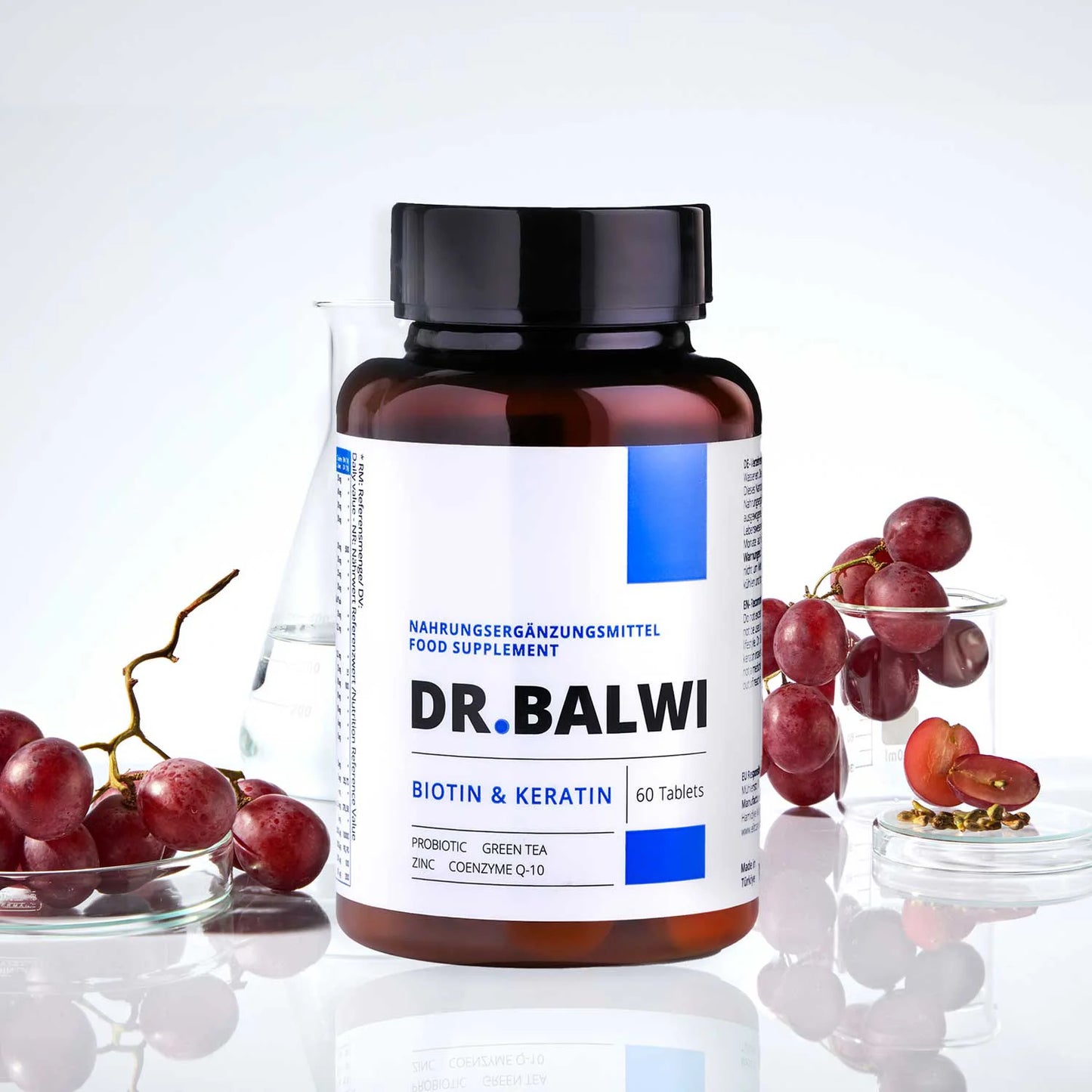 Eine Flasche Biotin- und Keratintabletten von shop.elithairtransplant zur Unterstützung des Haarwachstums ist auf einer weißen Fläche abgebildet. Daneben stehen rote Trauben, ein Becherglas und ein Glas mit klarer Flüssigkeit.