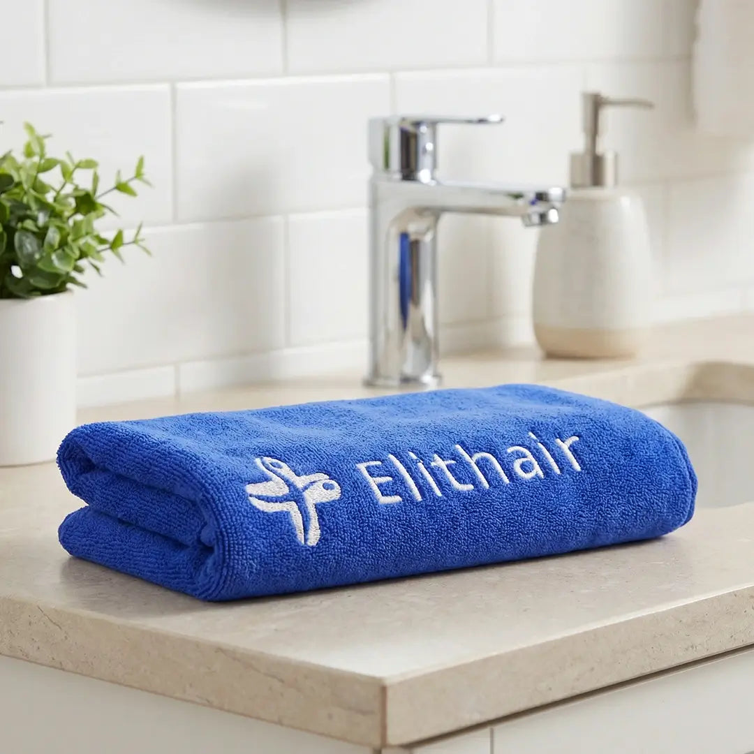 Ein blaues Mikrofaser-Kopfhandtuch aus dem Dr. Balwi Shop, bestickt mit "Elithair" und einem Logo, liegt auf einem Badezimmertisch in der Nähe eines Waschbeckens, mit einer Topfpflanze und einem Seifenspender im Hintergrund.