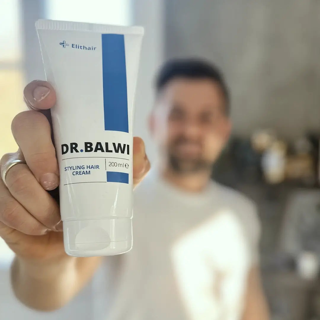 Eine Person hält eine Tube der Dr. Balwi Styling & Repair Creme aus dem Dr. Balwi Shop in der Hand und konzentriert sich auf das Produkt für glattes Haar, wobei der Hintergrund leicht verschwommen ist.