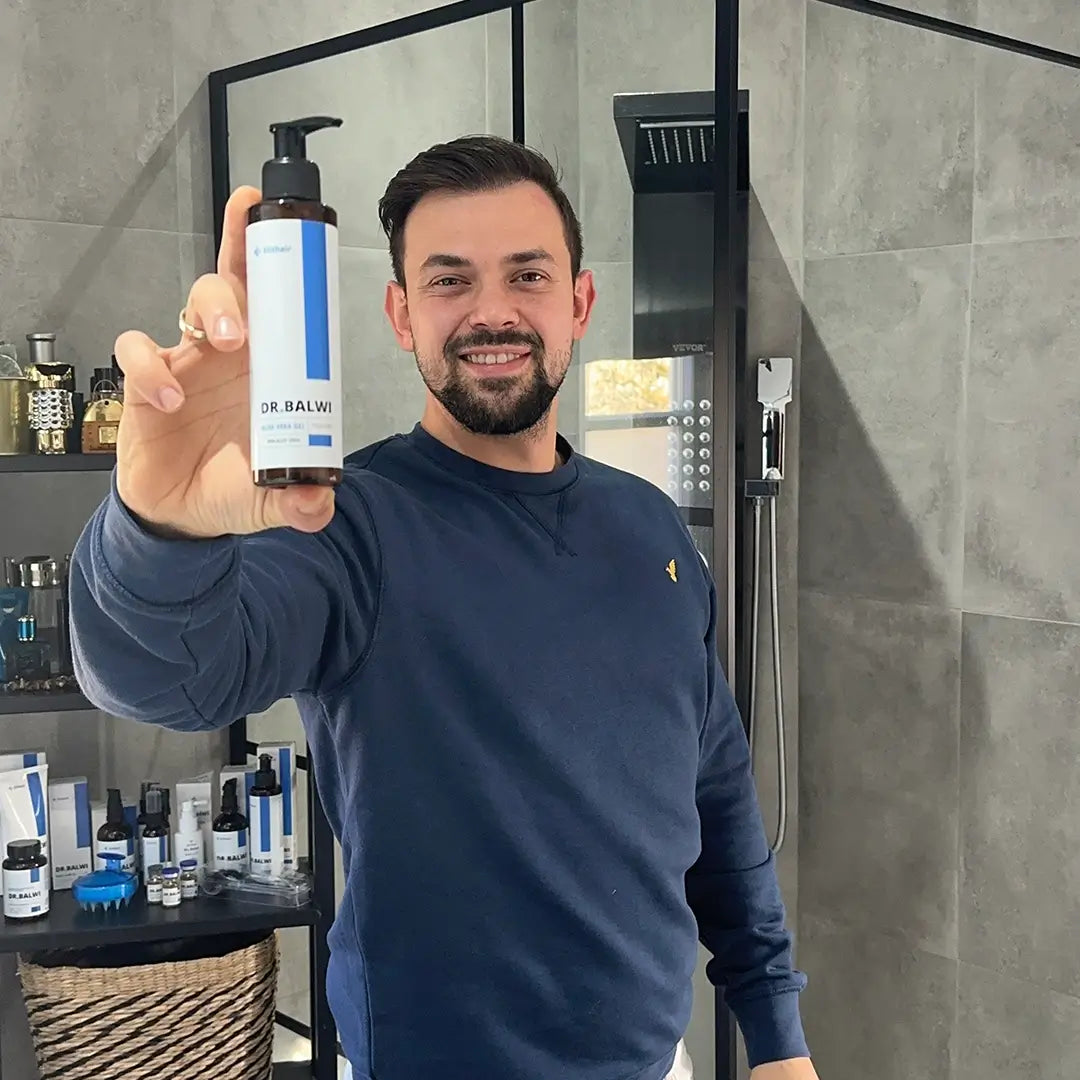 Ein bärtiger Mann in einem marineblauen Sweatshirt lächelt, als er eine Flasche Dr. Balwi 98% Aloe Vera aus dem Dr. Balwi Shop, ideal für Feuchtigkeitspflege Haare, in einem modernen Badezimmer mit grauen Fliesen und Toilettenartikeln auf dem Tresen hochhält.