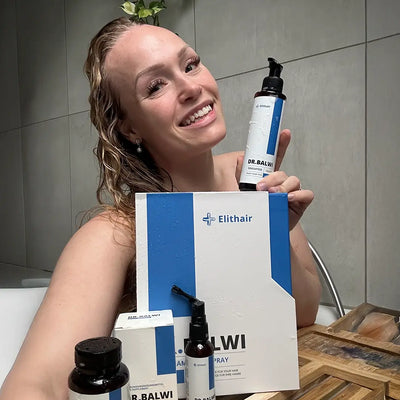 Eine lächelnde Frau mit nassem Haar hält eine Flasche aus dem Dr. Balwi Shop und sitzt hinter dem Anti-Haarausfall-Routine-Set auf einem hölzernen Badetablett im Badezimmer.