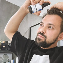 Ein bärtiger Mann sprüht sich Premium Schwarzkümmelöl von Dr. Balwi Shop auf die Haare in einem modernen Badezimmer, das die Haarpflege hervorhebt, wobei im Hintergrund Regale und Pflanzen zu sehen sind.