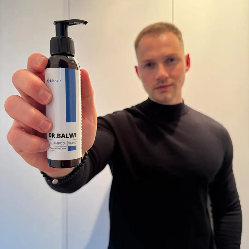 Ein Mann in einem schwarzen Rollkragenpullover hält das Dr. Balwi Shampoo aus dem Dr. Balwi Shop in die Höhe und hebt dessen Vorteile bei der Pflege von Haarausfall vor einem einfachen, verschwommenen Hintergrund hervor.