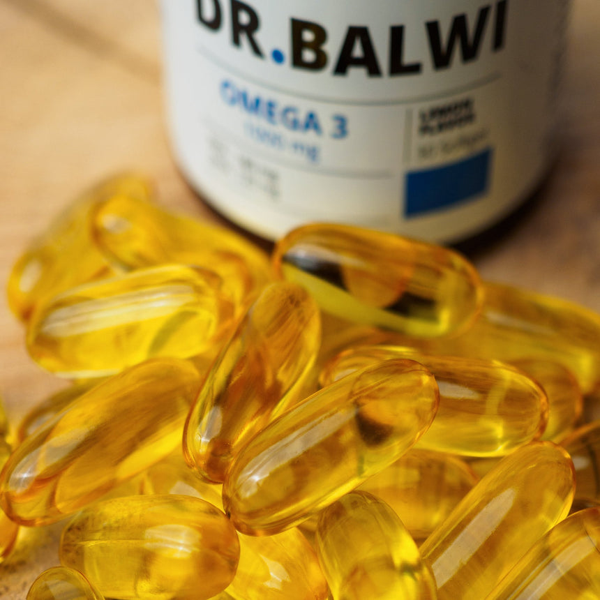 Ein Stapel gelber, durchsichtiger Daily Omega 3 Kapseln sitzt vor einer verschwommenen Dr. Balwi Shop Ergänzungsflasche, die Haarwachstum und gesunde Haarroutinen unterstützt.