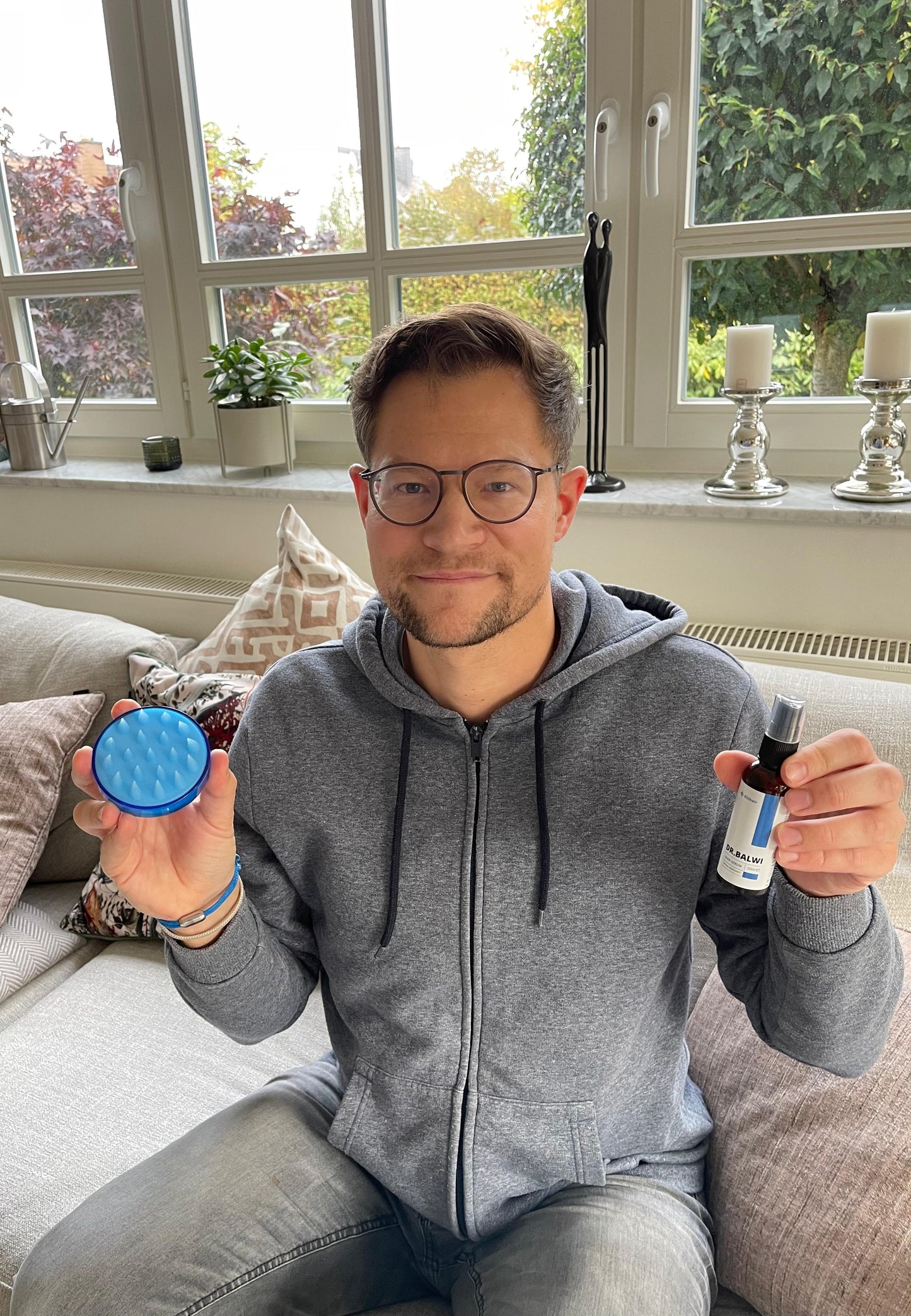 Ein Mann mit Brille und grauem Kapuzenpullover sitzt lächelnd auf einem Sofa und hält ein blaues Kopfhautmassagegerät und eine Flasche Dr. Balwi Haarserum von shop.elithairtransplant in der Hand. Hinter ihm ist ein helles Fenster mit Pflanzen und Kerzen.